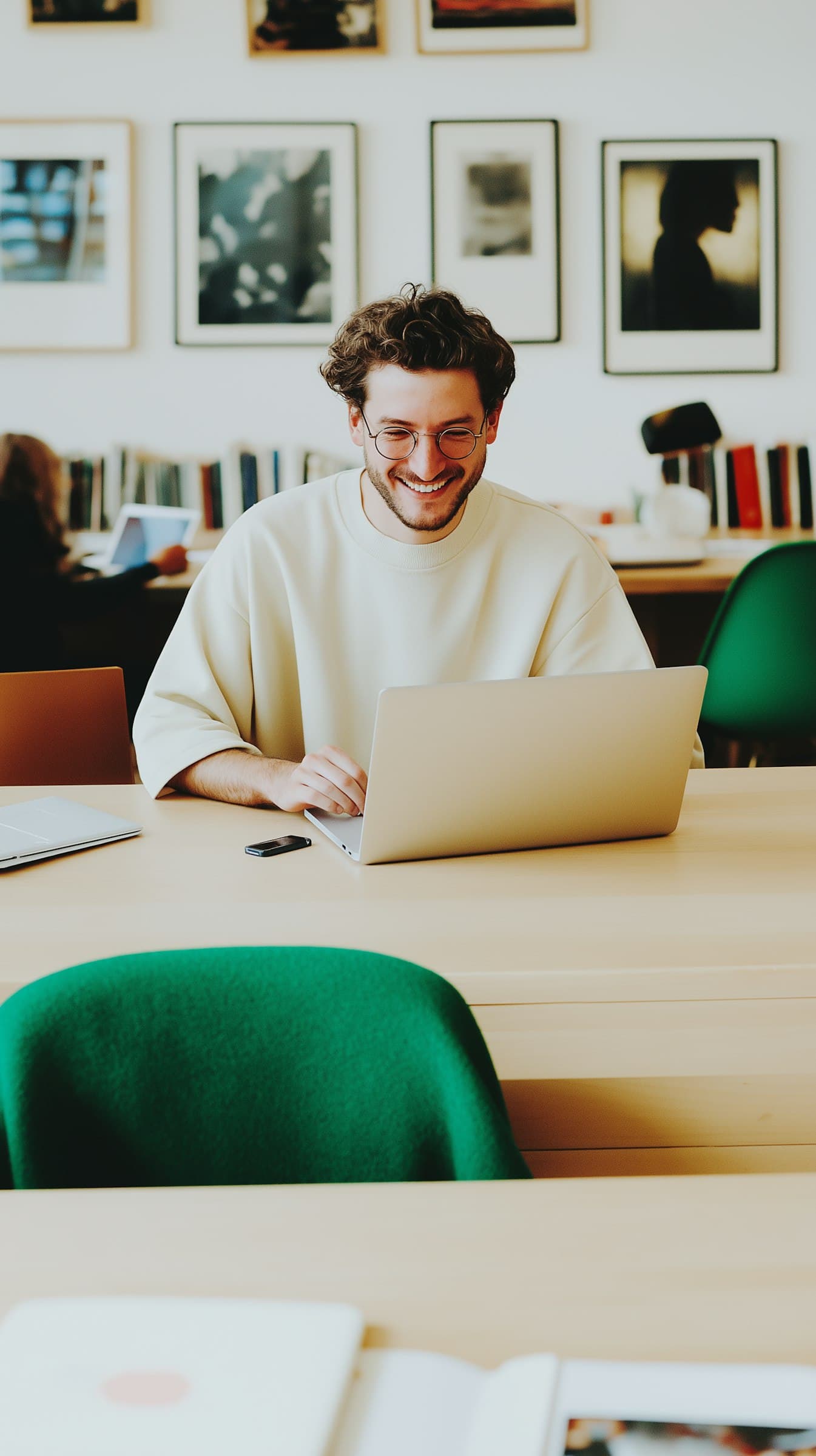 Smiling person using a laptop in a library with green chairs and framed art on the wall.